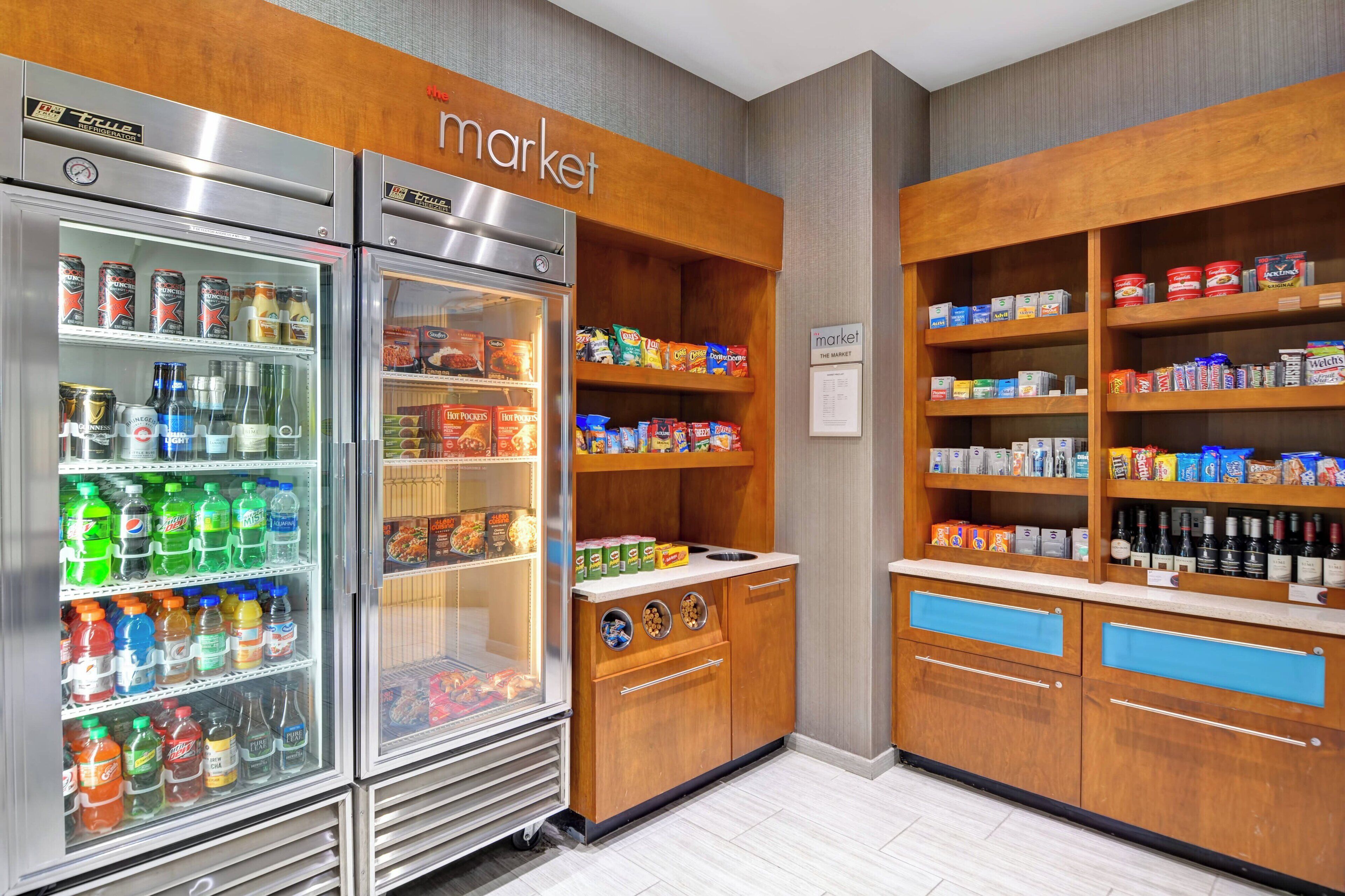 Una kitchenette con refrigerador lleno de bebidas y aperitivos, un mueble de madera con estantes de alimentos, y un letrero que indica un área de mercado.