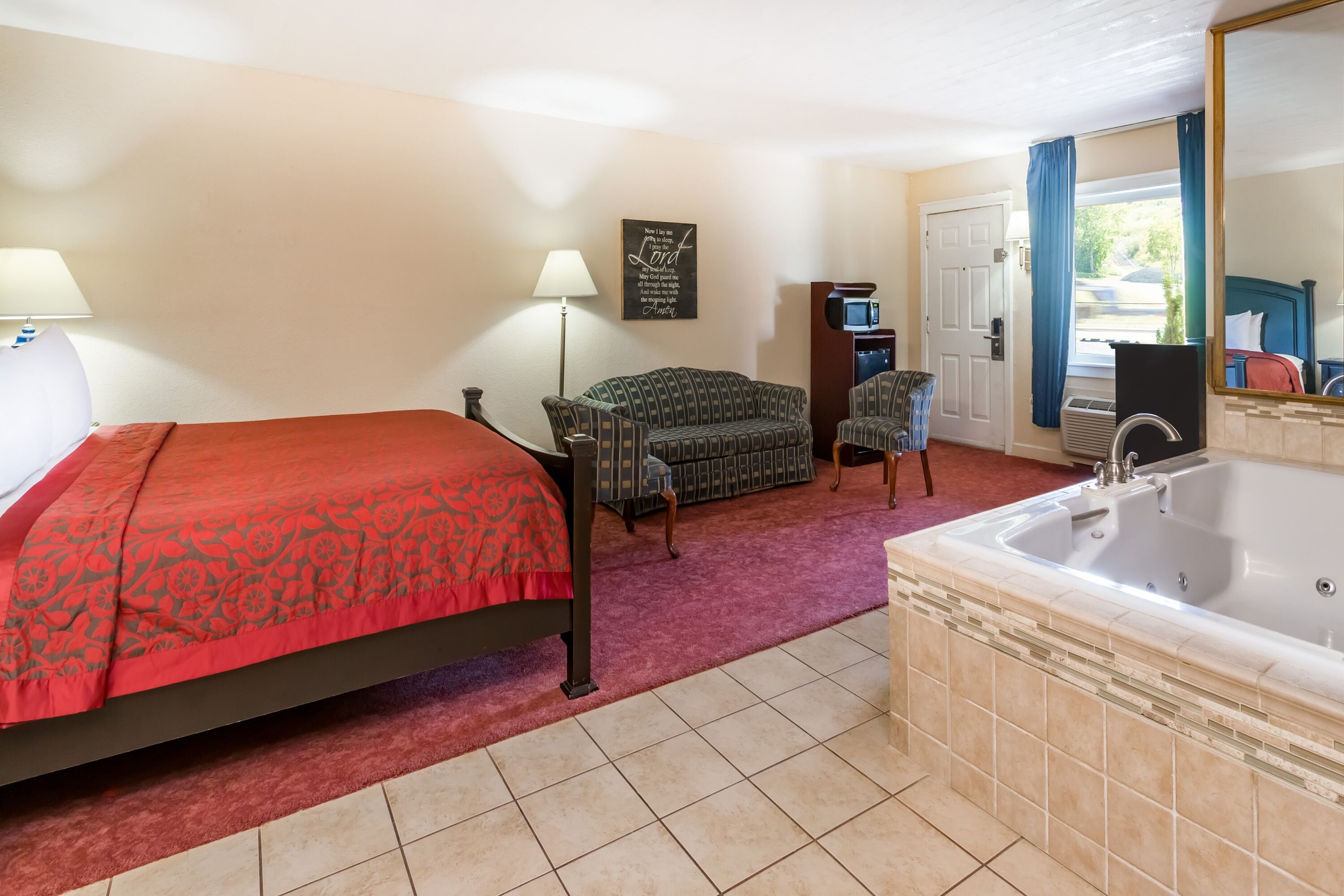 Habitación de hotel con cama, sofá, una mesita, una silla, baño con bañera y ventana con cortinas.