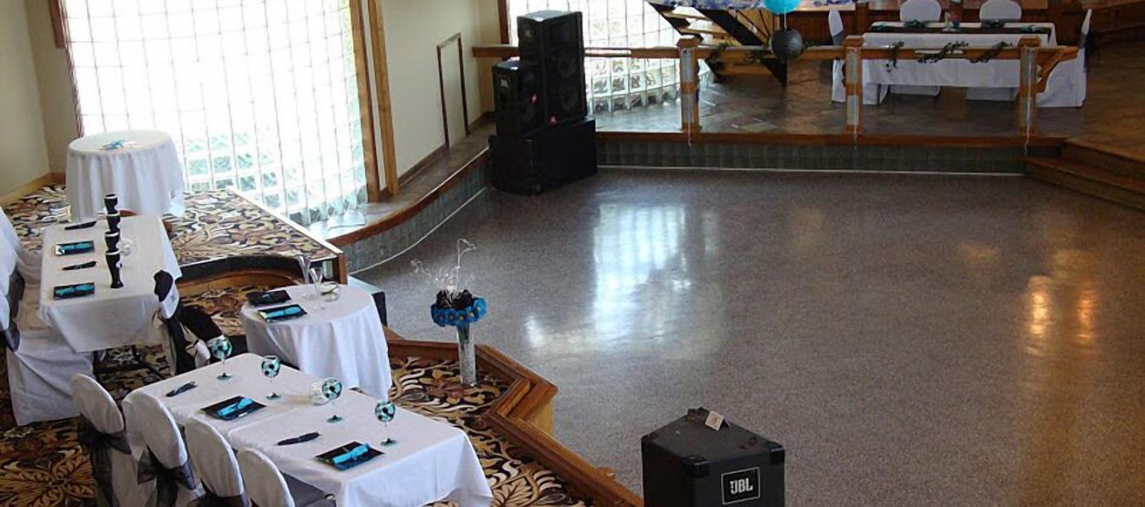 A banquet hall with round tables set for a formal event, a stage with a speaker system, and a view of the seating area through a glass partition.