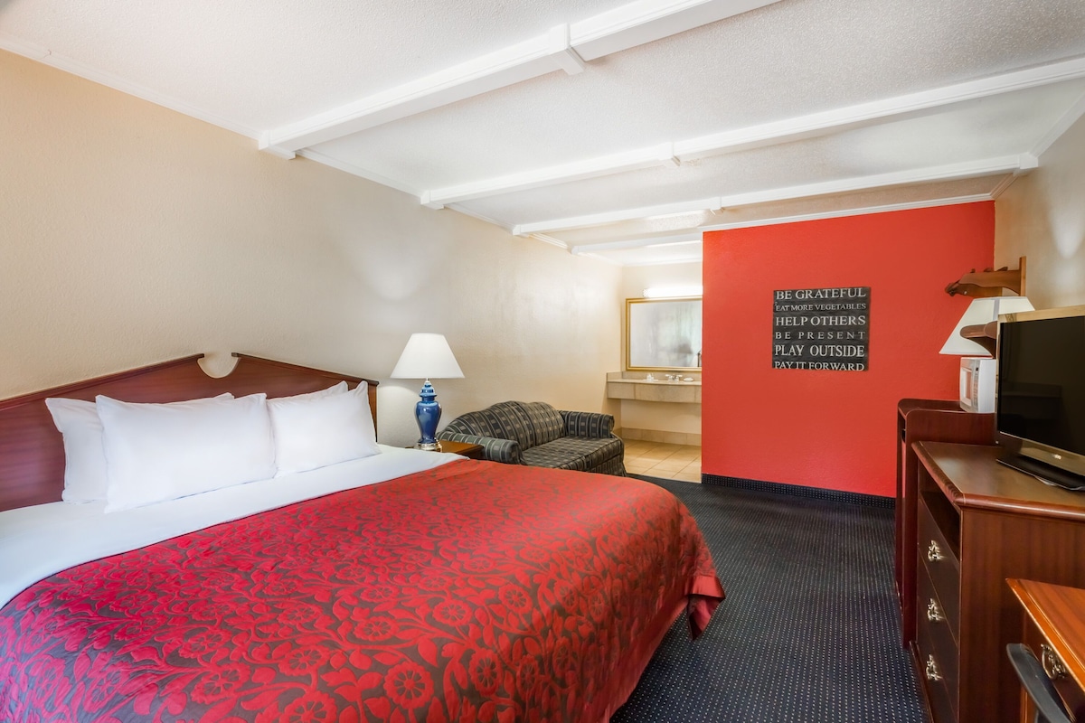 A hotel room with a large bed, a television, a desk with a lamp, a chair, and a motivational quote on the wall.