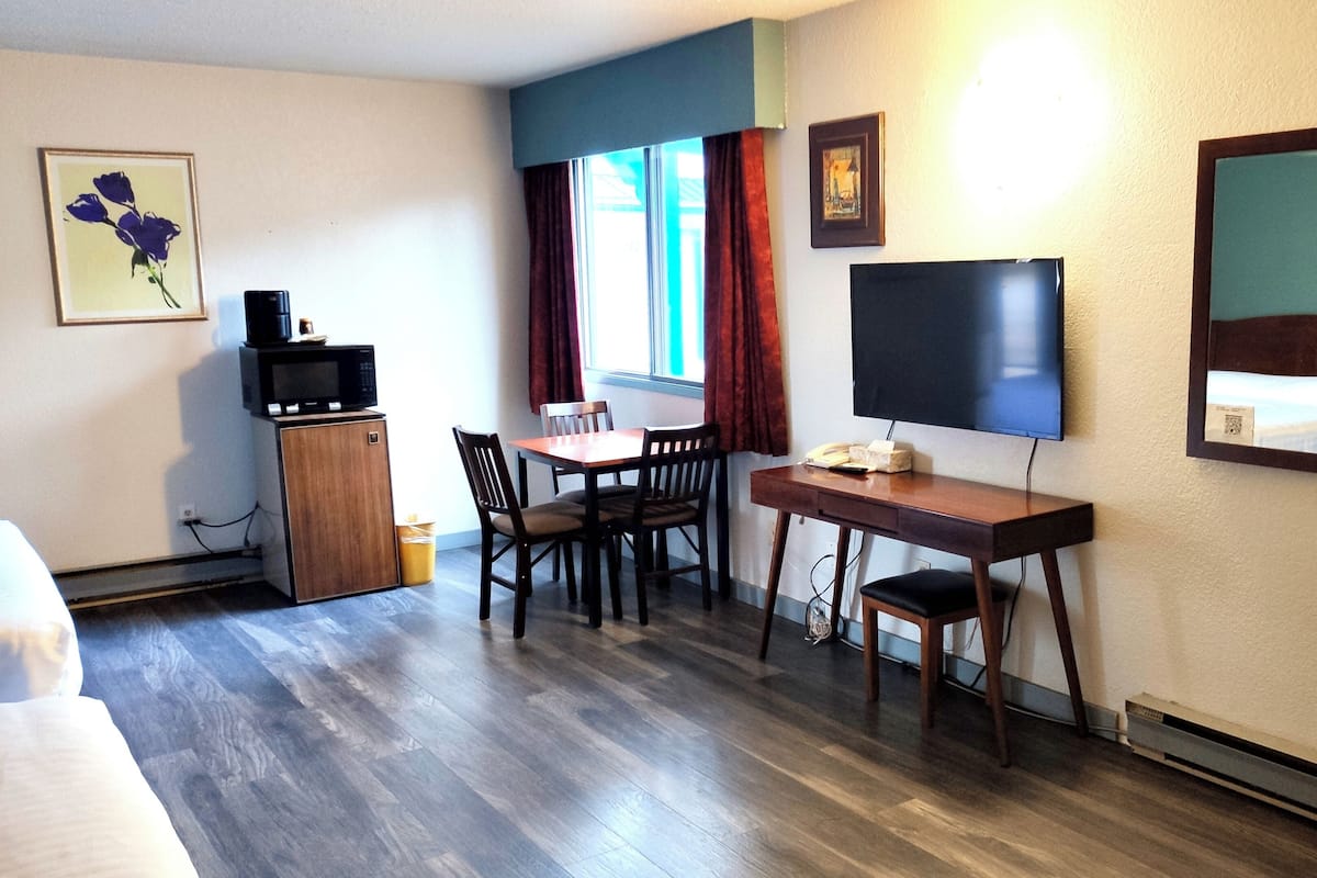 A hotel room with a wooden floor, a desk with a chair, a television, a small cabinet with a microwave, and a window with curtains.