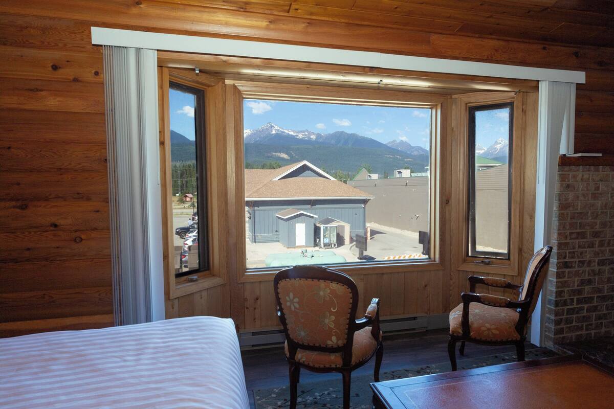 A room with a large window offering a view of mountains and a building. There are two wooden chairs and a bed.
