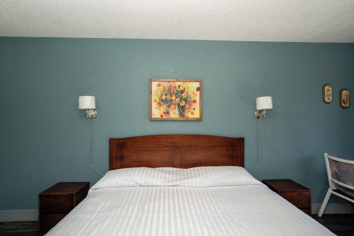 A bedroom with a wooden headboard, a bed with white bedding, two bedside tables, a nightstand, a chair, and a wall-mounted painting.