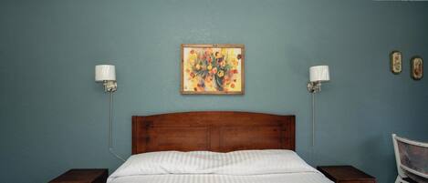 A bedroom with a wooden headboard, a bed with white bedding, two bedside tables, a nightstand, a chair, and a wall-mounted painting.