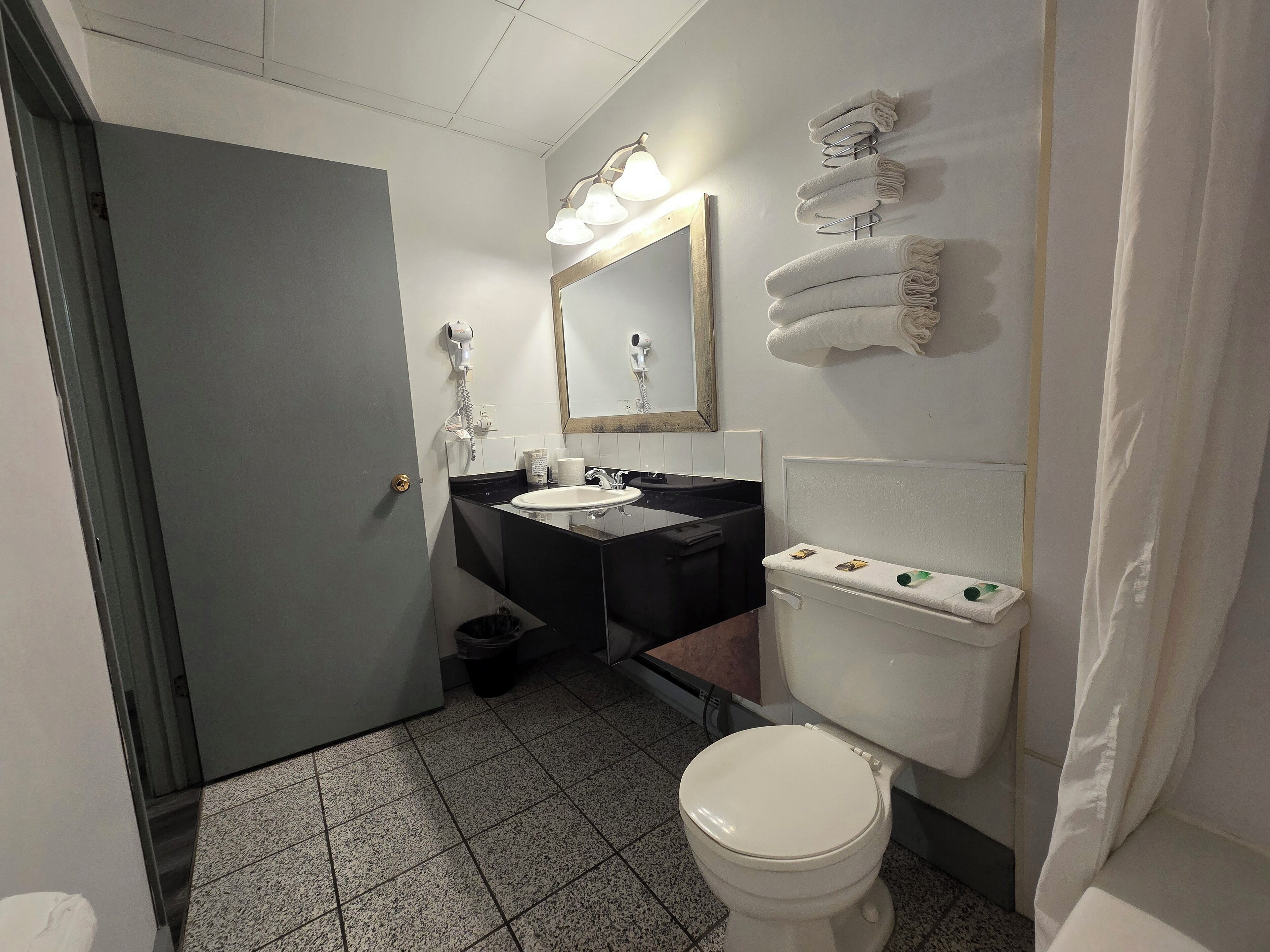 Un baño con inodoro, lavamanos y espejo. Hay toallas en la pared y un secador de pelo también en la pared.