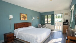 A bedroom with a large bed, bedside tables, a desk, and a chair. There is a window with curtains, a painting on the wall, and a view of the outside. - Alpine Inn (Valemount)