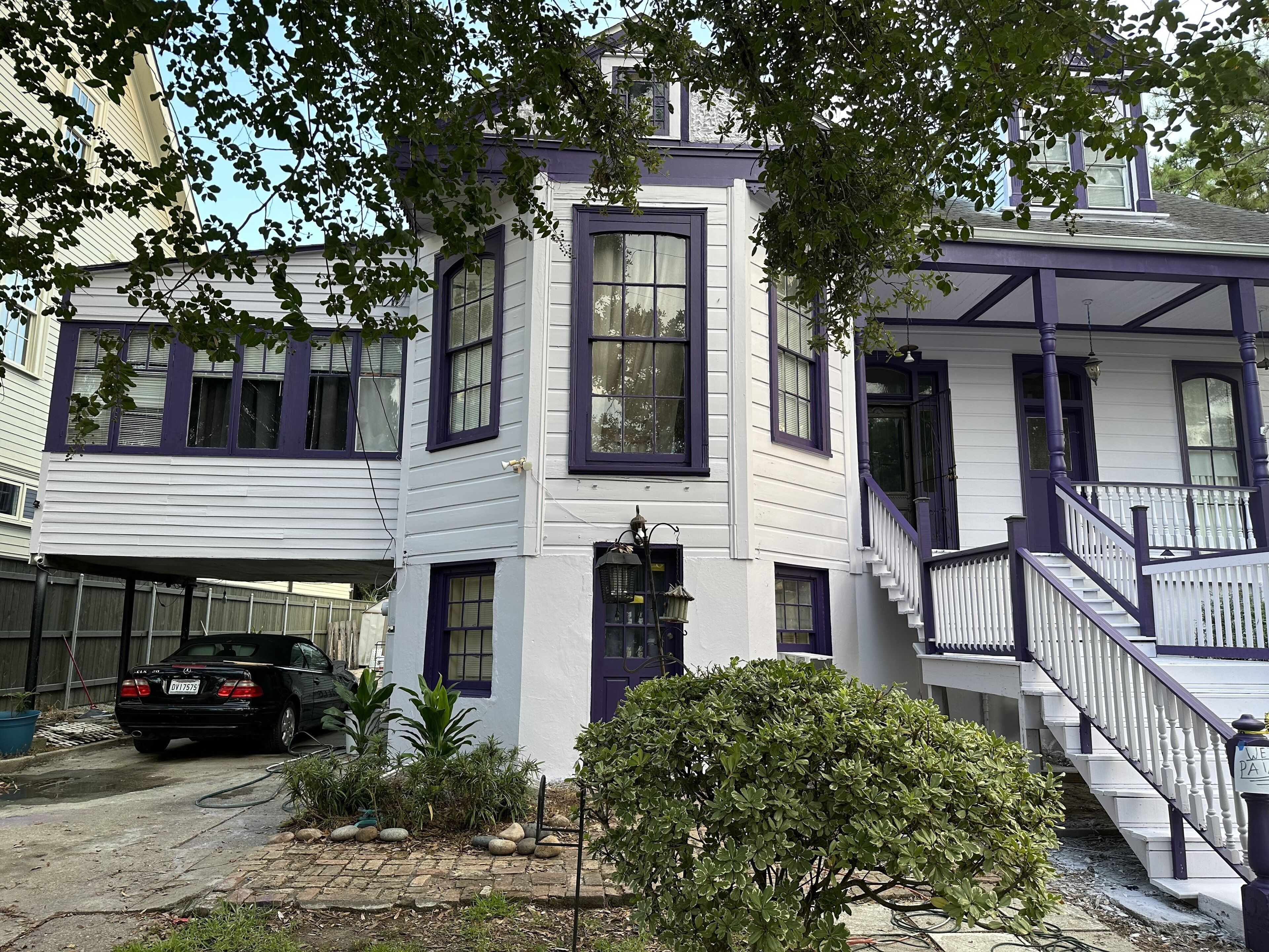 A two-story house with a white exterior and purple trim. There is a black car parked in the driveway, and a small garden with various plants and rocks.