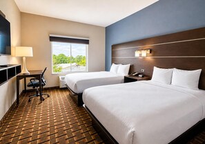 A hotel room with two beds, a desk with a lamp, a TV, and a window with a view of trees.