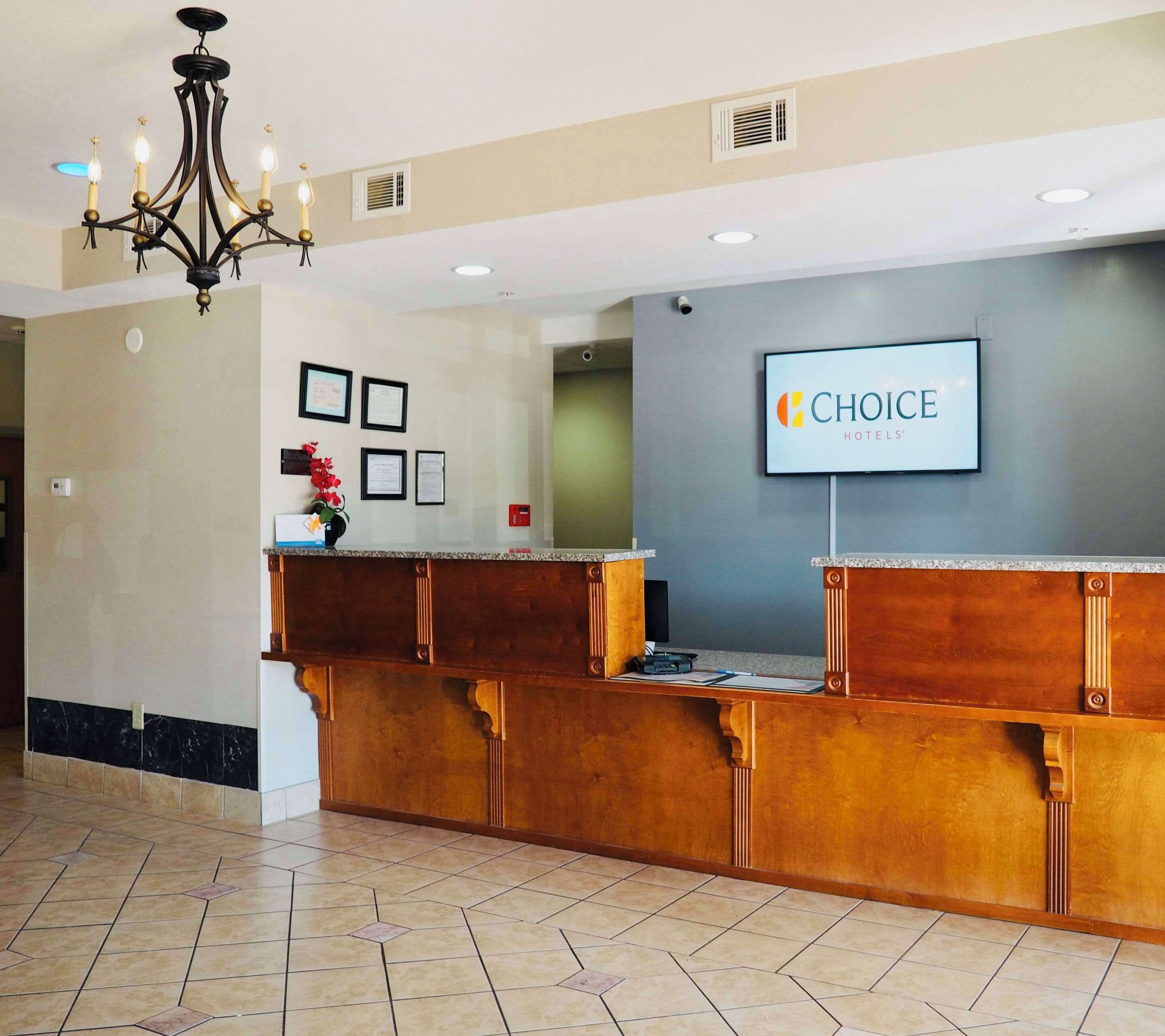 A reception area with a wooden reception desk, a chandelier, framed certificates on the wall, and a television displaying the hotel name.