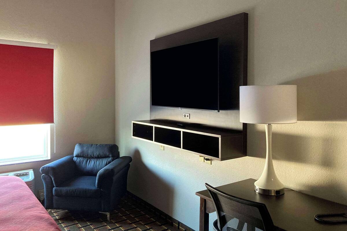 A hotel room with a flat-screen TV mounted on the wall, a desk with a lamp, a chair, and a bed with a red headboard.