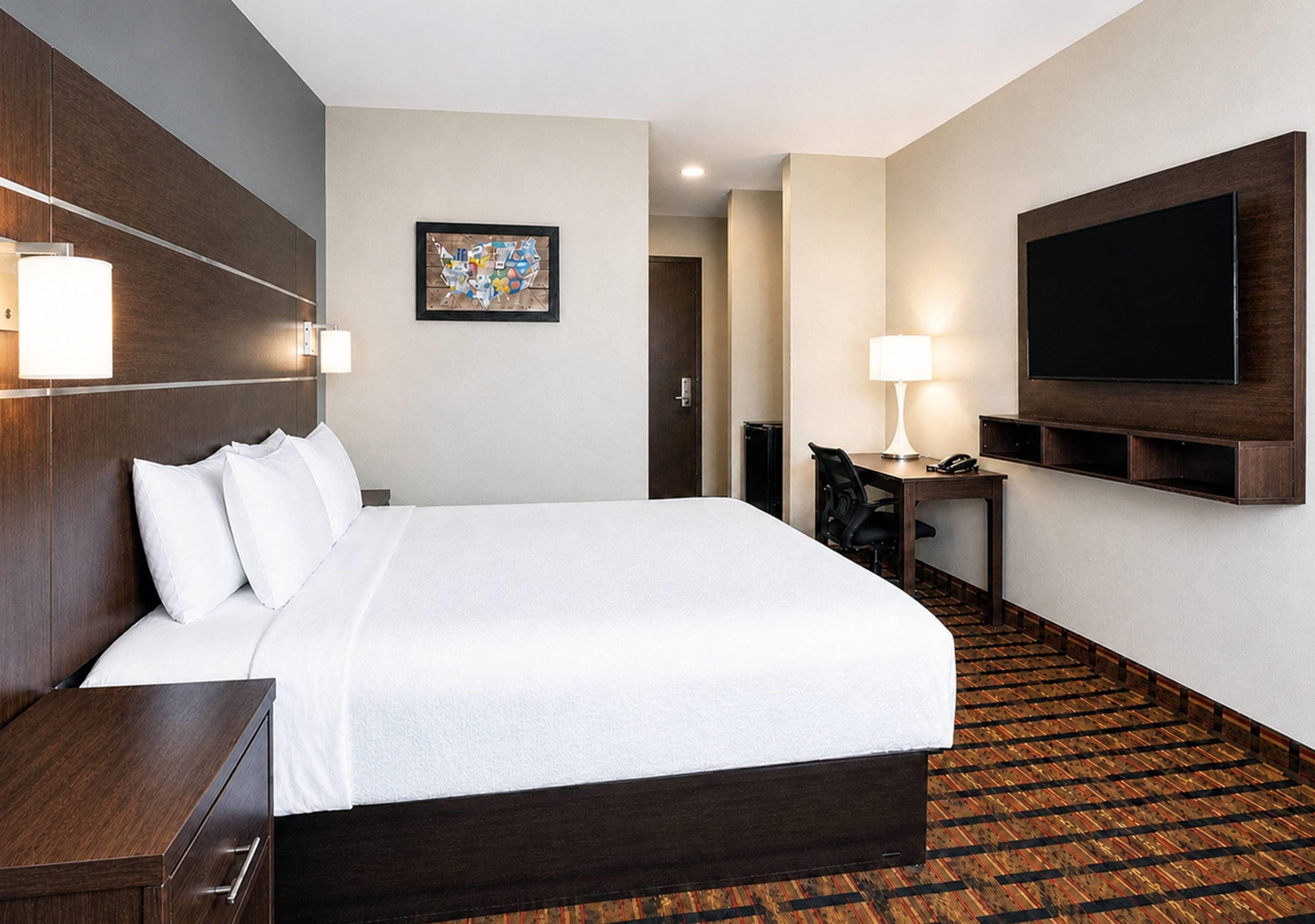A hotel room with a large bed, bedside tables, a desk with a chair, a flat-screen TV mounted on the wall, and a patterned carpet.