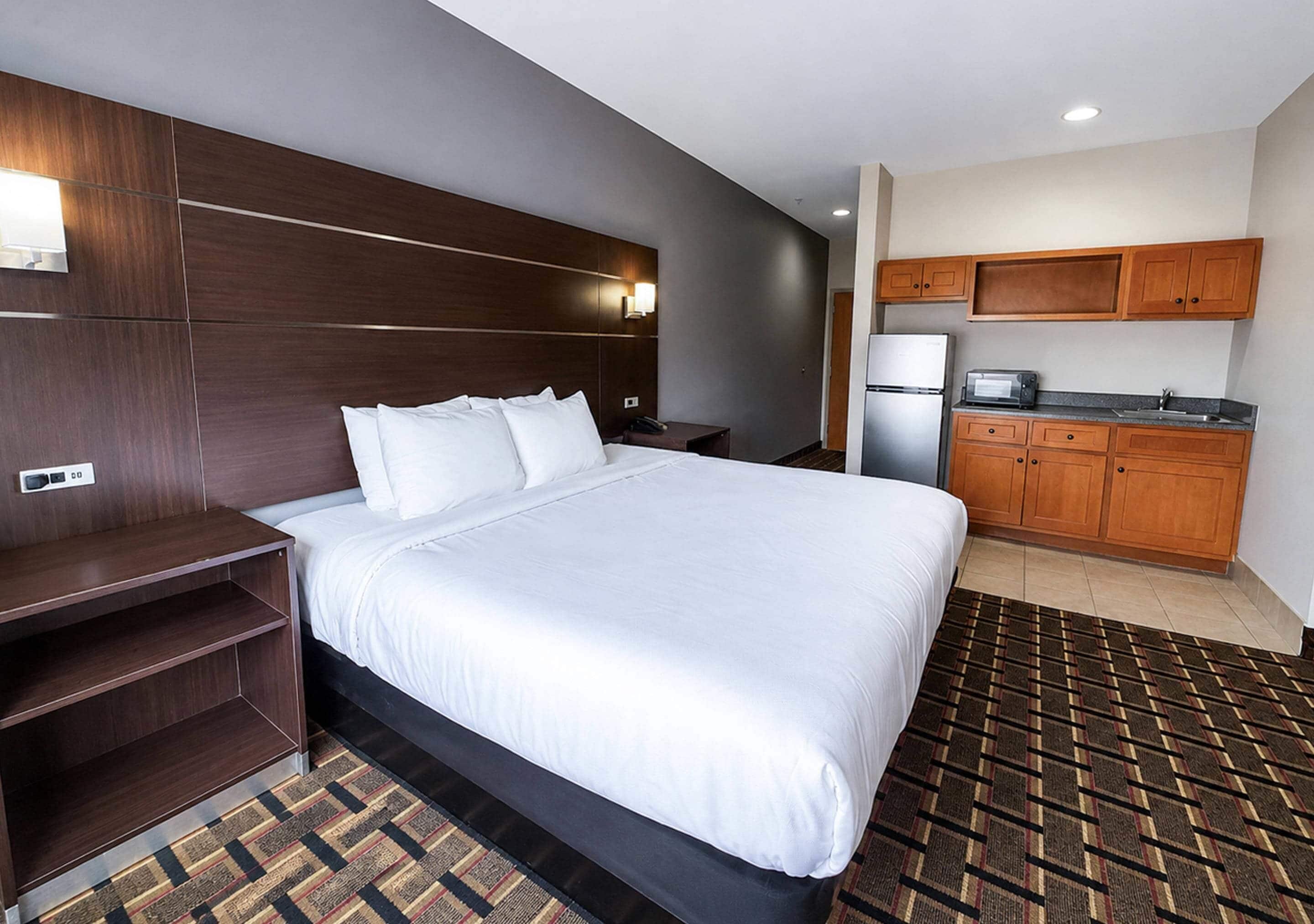 A hotel room with a large bed, a wooden headboard, a small kitchenette, and a patterned rug.