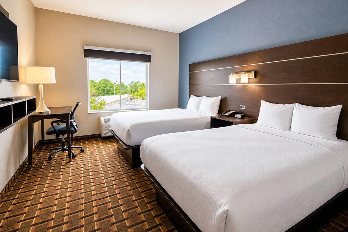 Habitación de hotel con dos camas, un escritorio con lámpara, un televisor y una ventana con vista a árboles.