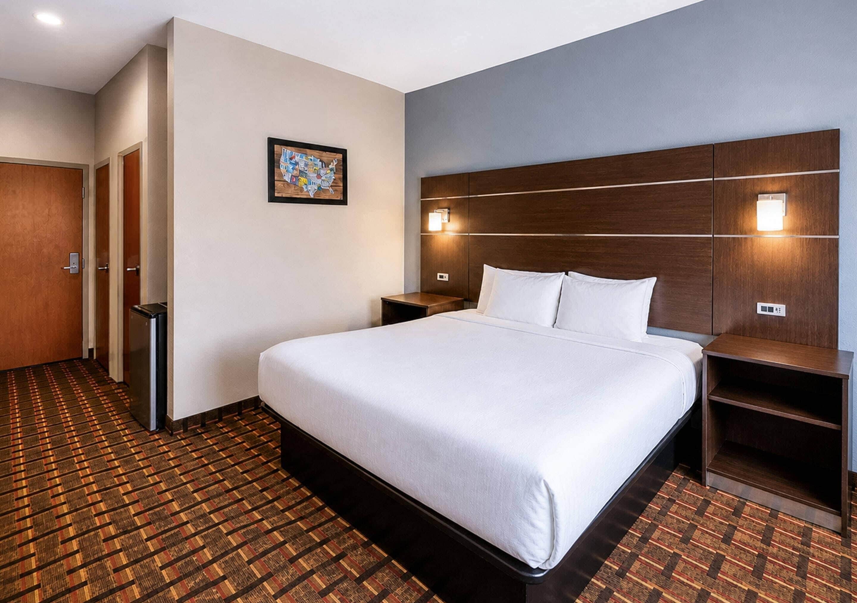 A hotel room with a large bed, a headboard with integrated lighting, a small bedside table, a nightstand, and a patterned carpet.