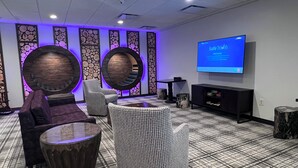 A modern lounge area with a flat-screen TV displaying 'Safe Travels Welcome!' on the wall, a round wooden coffee table, two armchairs, and a patterned ottoman.
