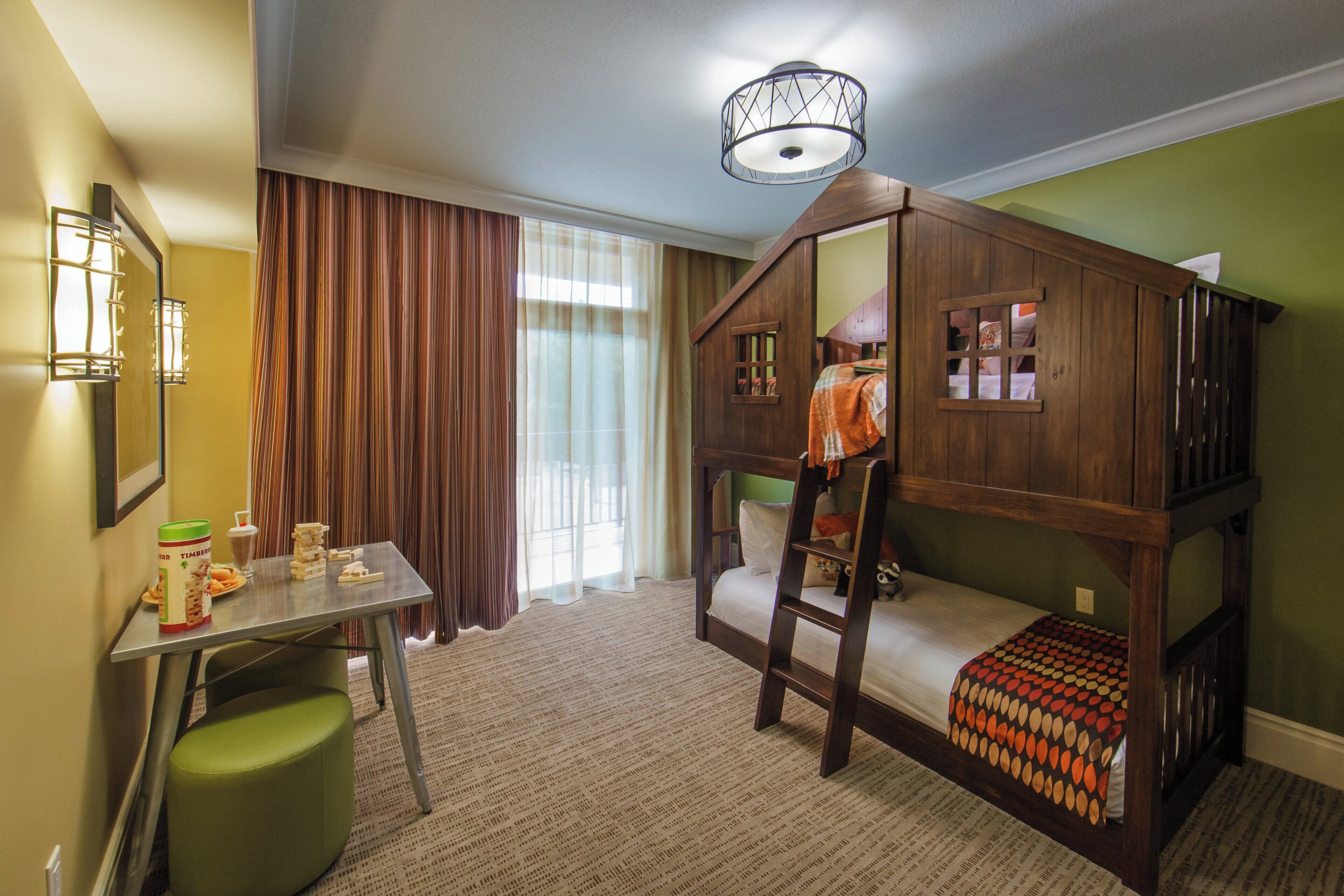 A child's bunk bed with a ladder, a desk with a chair, a window with curtains, and a wall-mounted lamp.