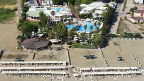 An aerial view of a coastal resort with multiple swimming pools, beach umbrellas, and a pier extending into the sea.