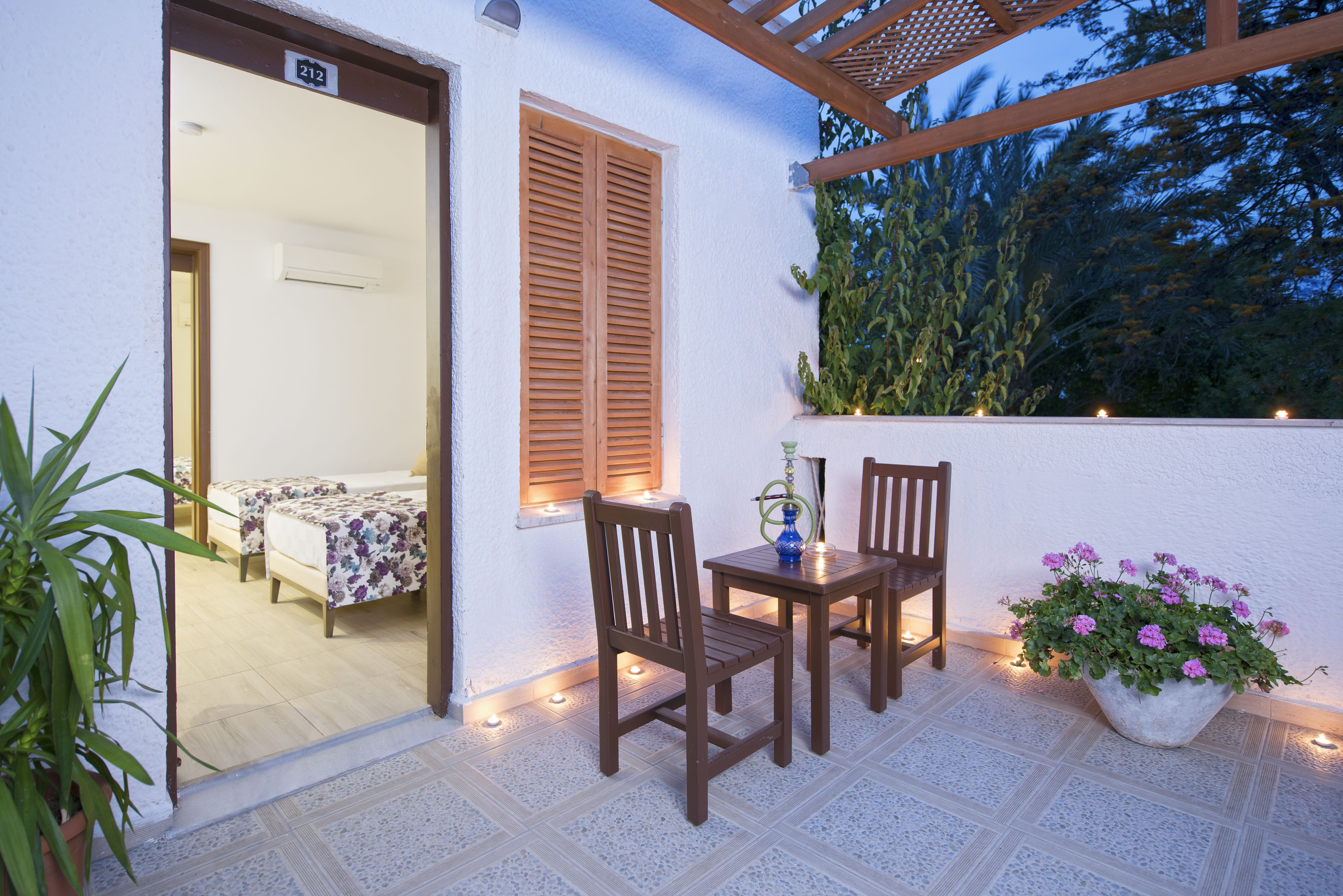 Eine Terrasse im Freien mit einem Holztisch und Stühlen, einem Blumentopf mit rosa Blüten und Blick auf ein Schlafzimmer durch eine Glastür.