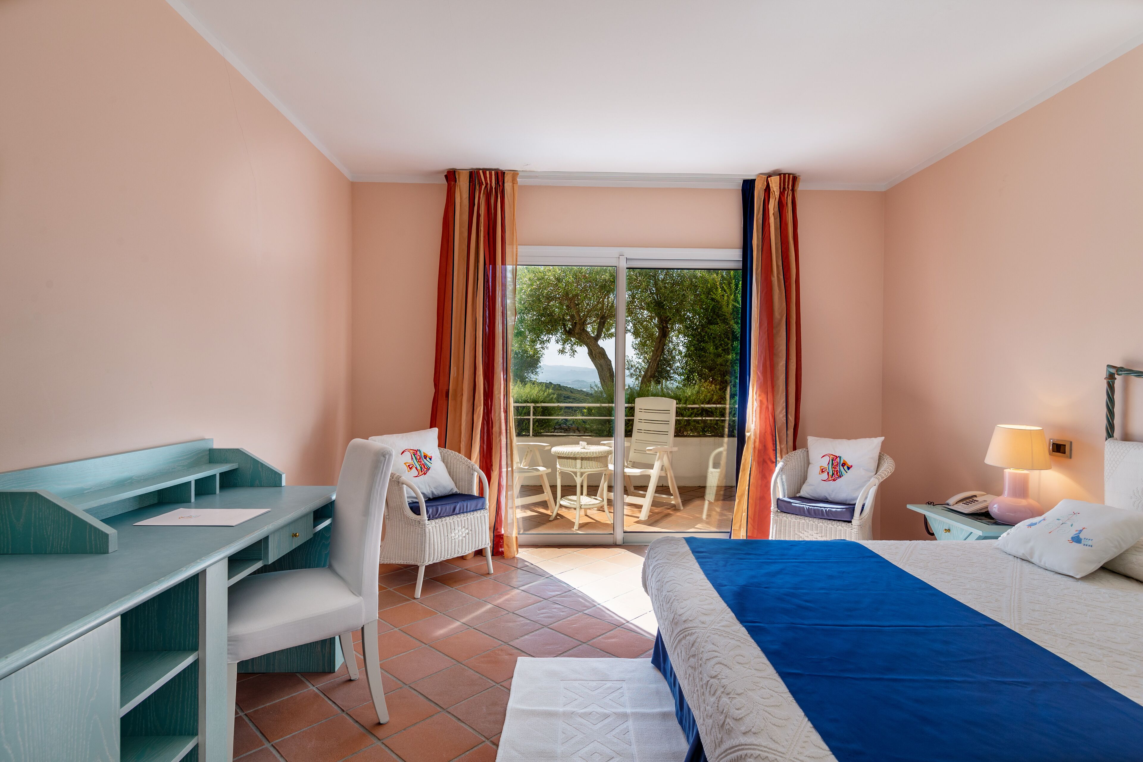 A hotel room with a bed, a desk, two chairs, a balcony with a table and chairs, and a view of trees and water.
