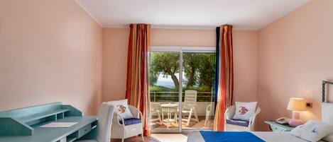 A hotel room with a bed, a desk, two chairs, a balcony with a table and chairs, and a view of trees and water.