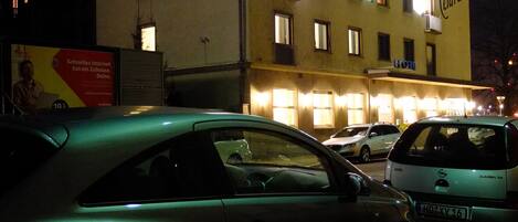 Edifício de hotel com vários andares, janelas iluminadas e varandas, com carros estacionados na frente.