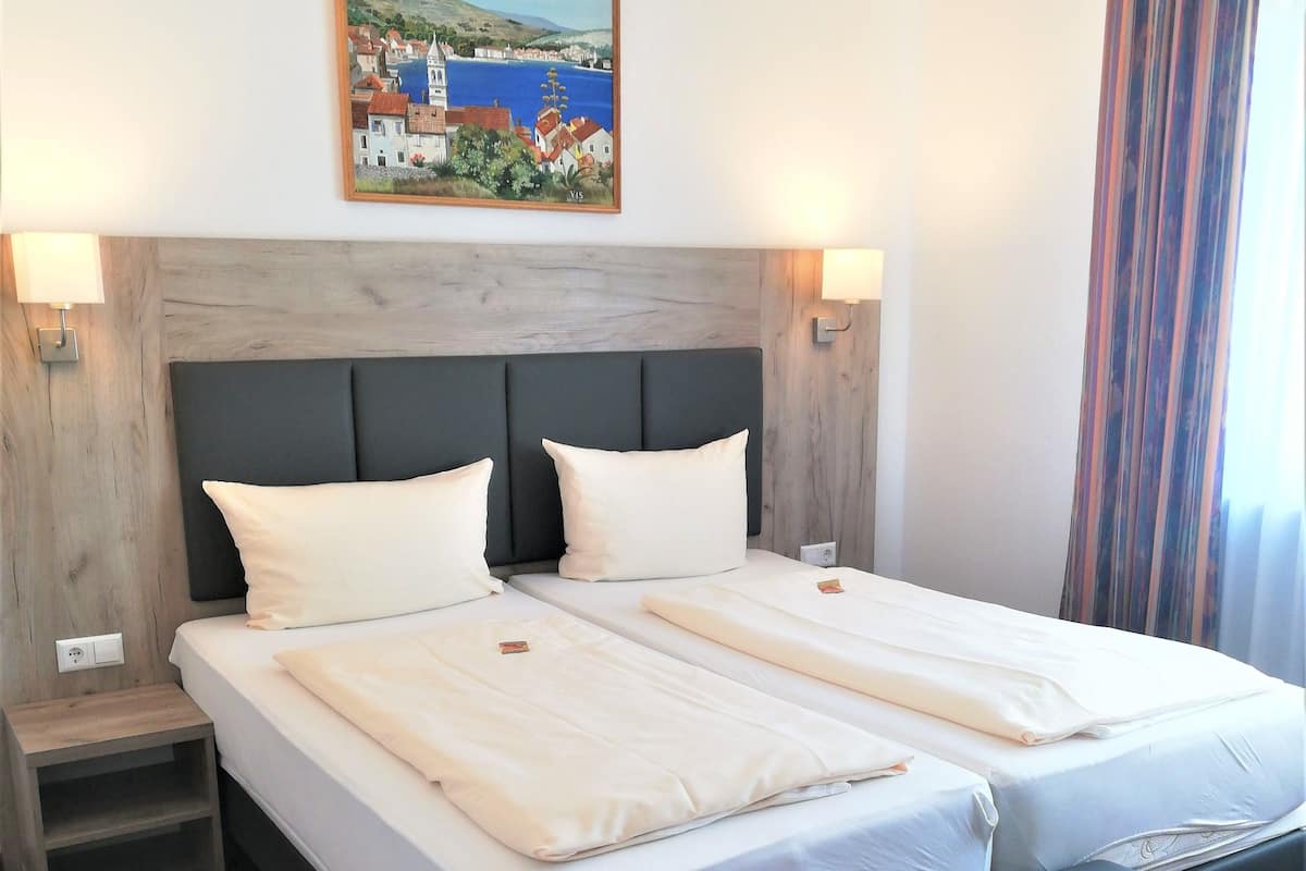 A double bed with a grey headboard, two bedside tables, a small wooden nightstand, a large window with striped curtains, and a painting of a coastal scene above the bed.