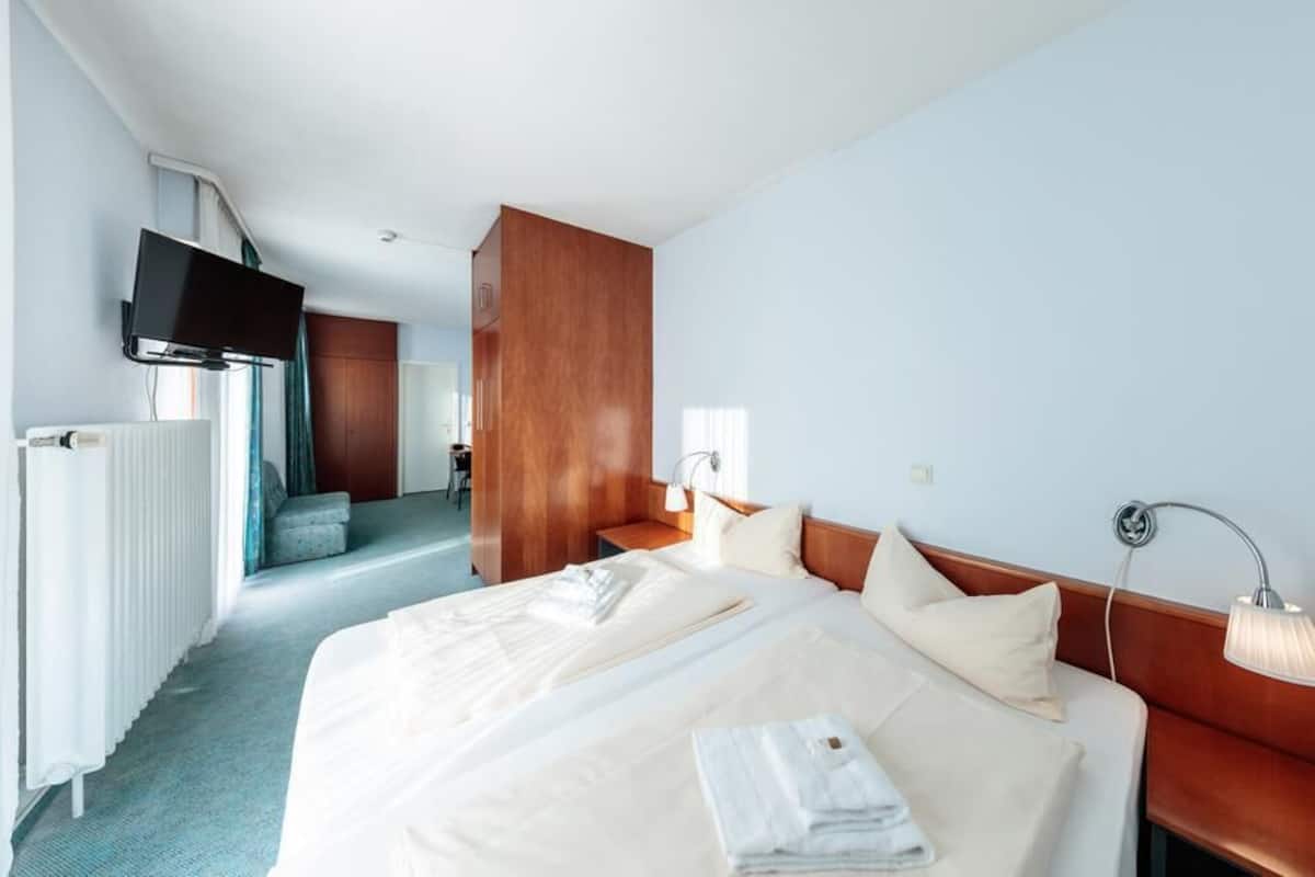 A hotel room with a large bed, a TV mounted on the wall, a wooden wardrobe, and a bathroom visible through an open door.
