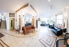 Een ruime woonkamer met een bruine bank, een glazen salontafel en een zwarte leren bank. Links bevindt zich een trap en aan de rechterkant een eethoek met een tafel en stoelen.