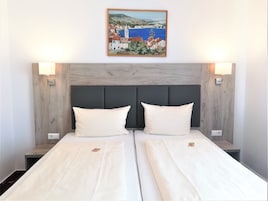 Een tweepersoonsbed met twee kussens, een houten hoofdbord en daarboven een schilderij van een kustplaatsje.