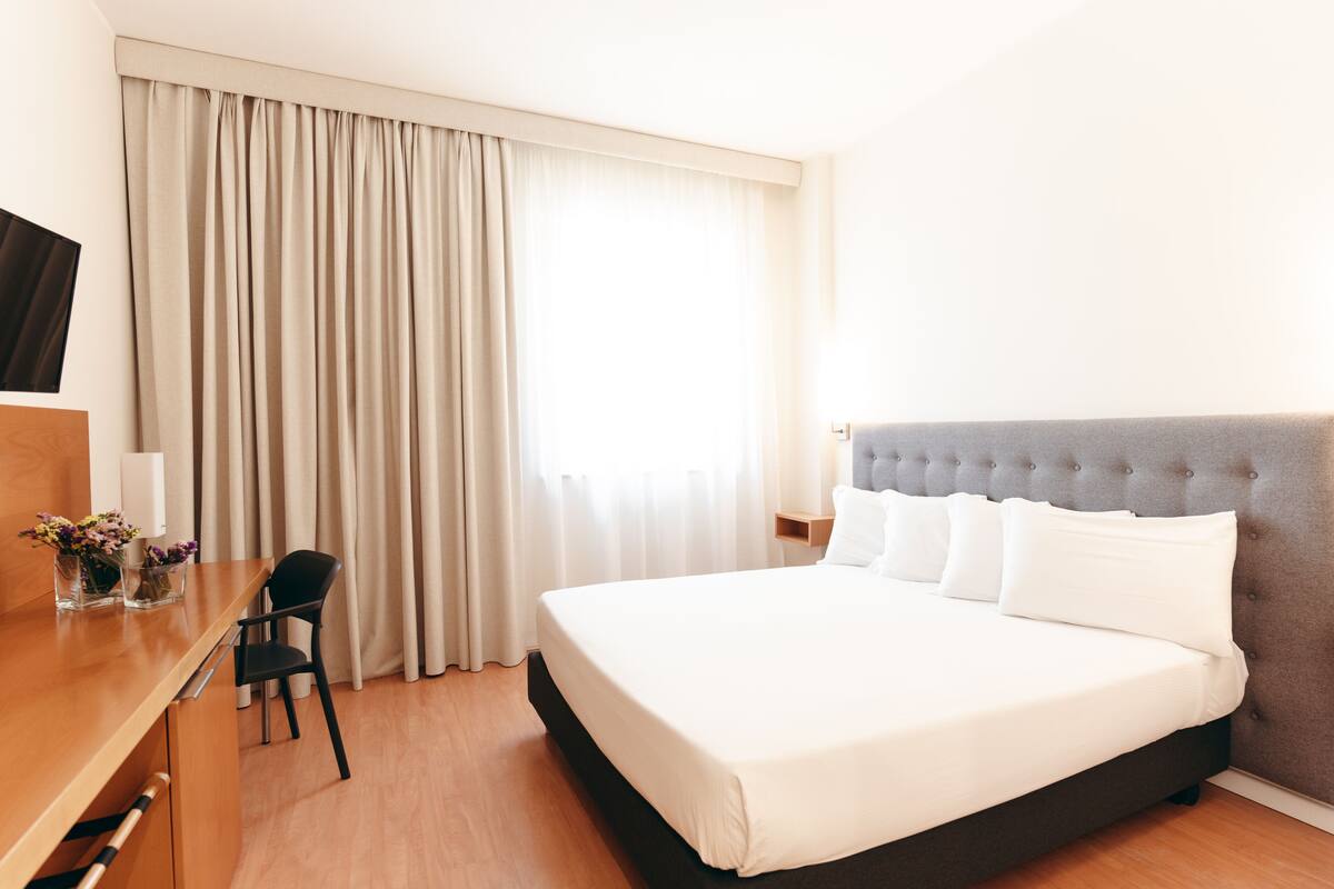 Ein Hotelzimmer mit Bett, Schreibtisch, Stuhl, Fernseher und einem Fenster mit Vorhängen.
