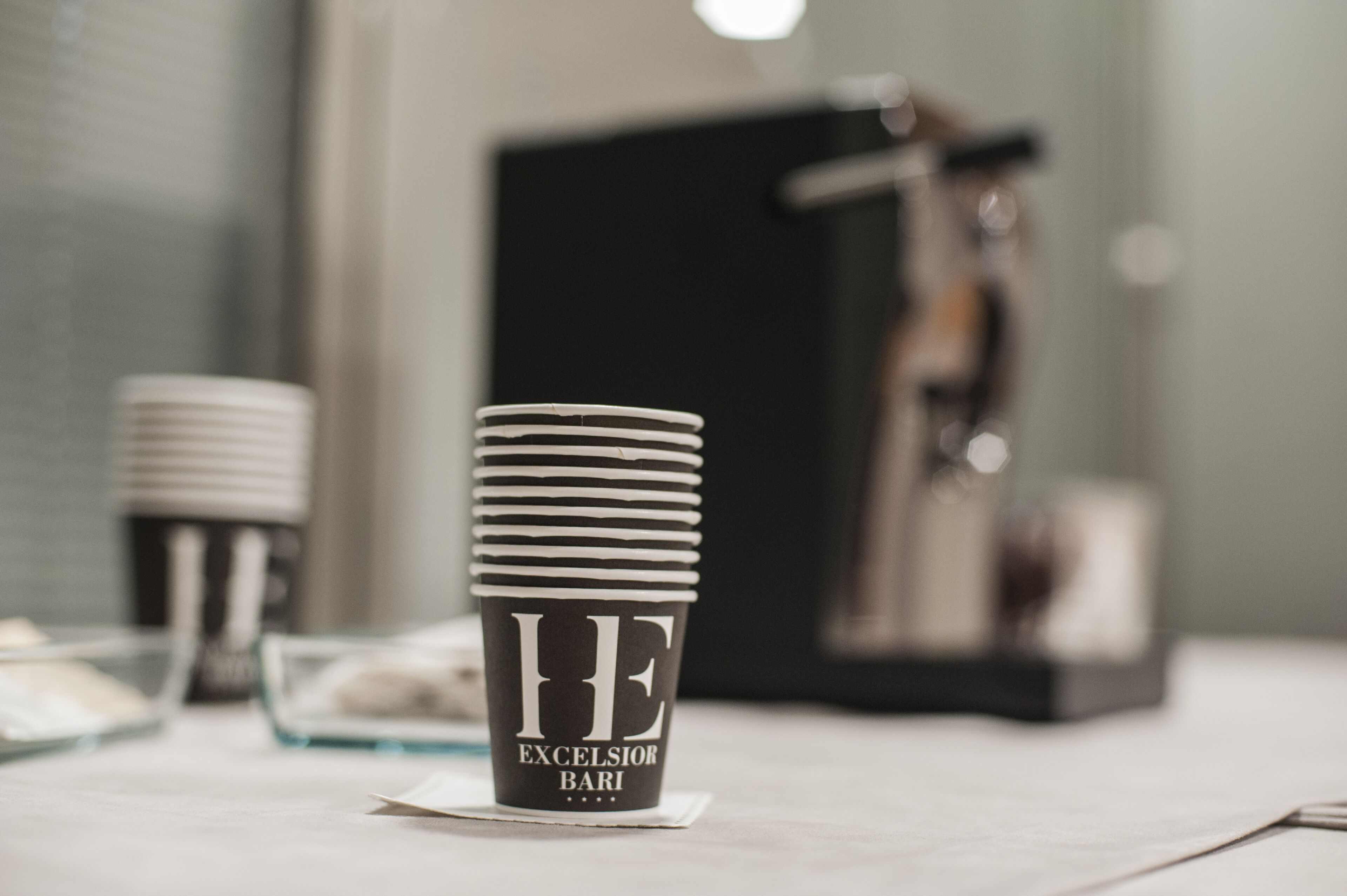 A stack of black and white cups with "HE EXCELSIOR BARI" printed on them, placed on a table with a glass container and a black box in the background.