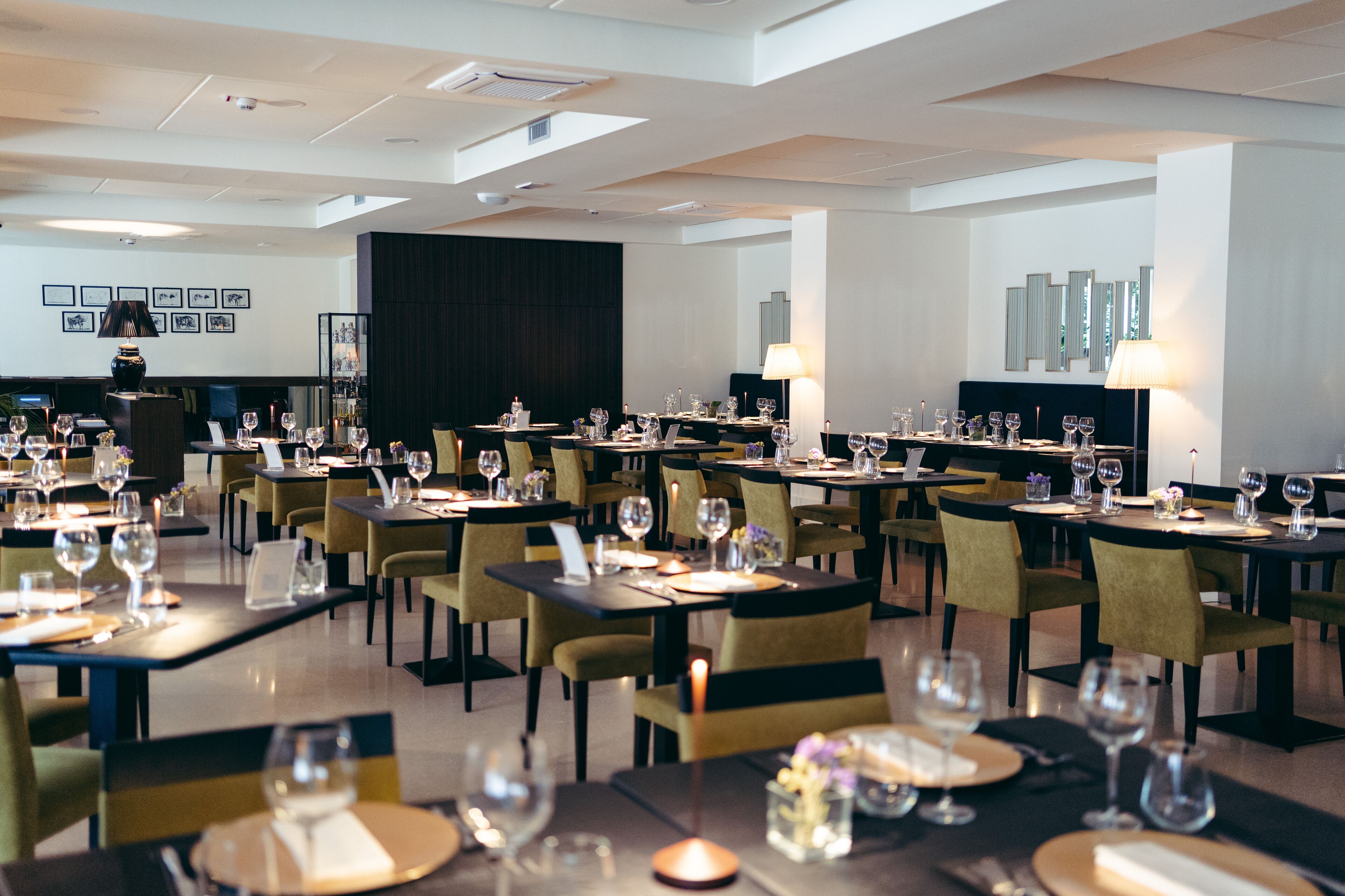 A modern dining area with multiple tables set for dining, featuring candles and wine glasses.