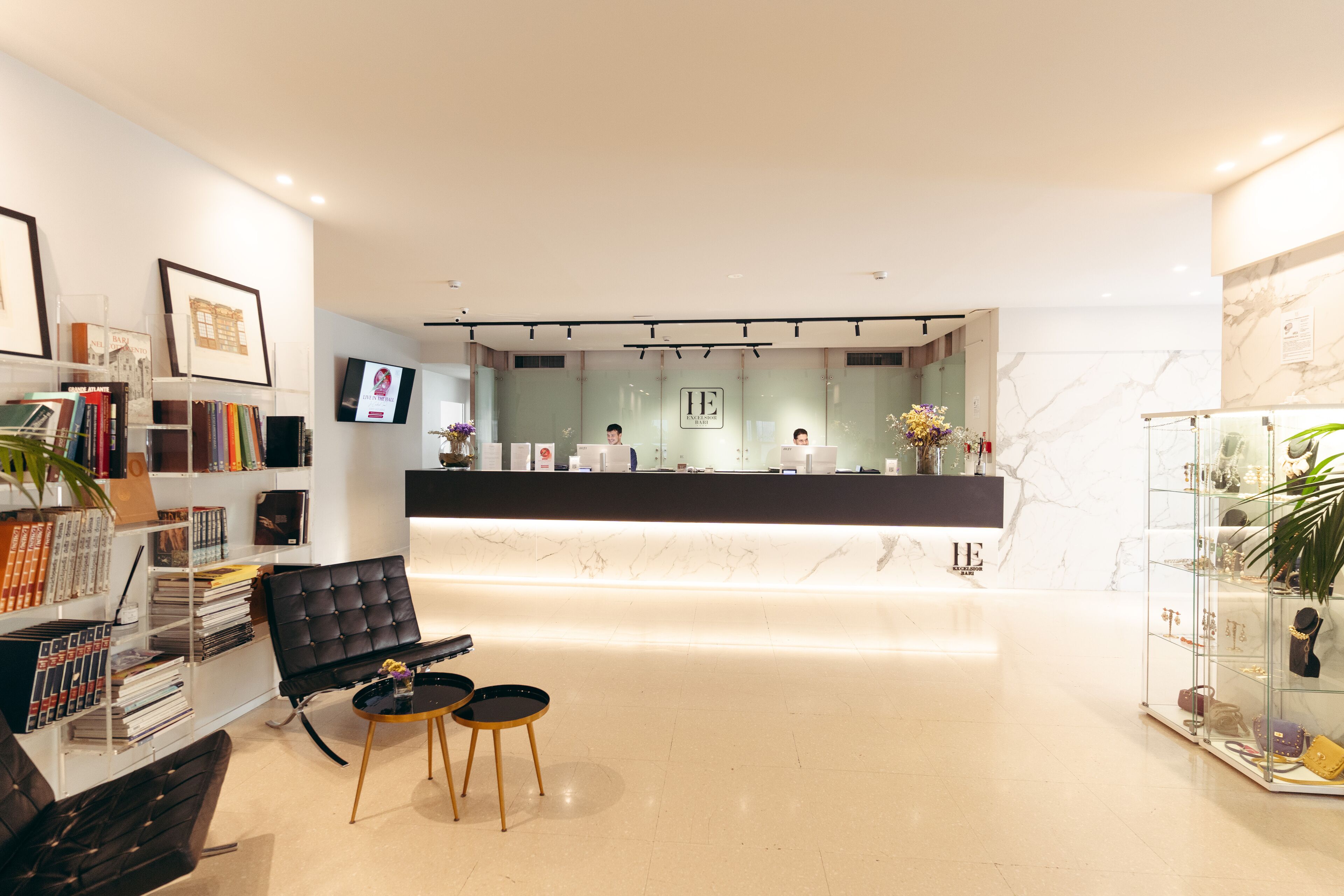 A modern hotel lobby with a reception desk, a bookshelf, a seating area, and a display of handbags.