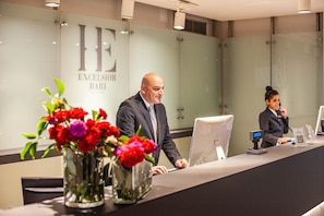 Un mostrador de recepción con un hombre y una mujer, un jarrón de flores y un cartel que dice 'Excelsior Bari'.