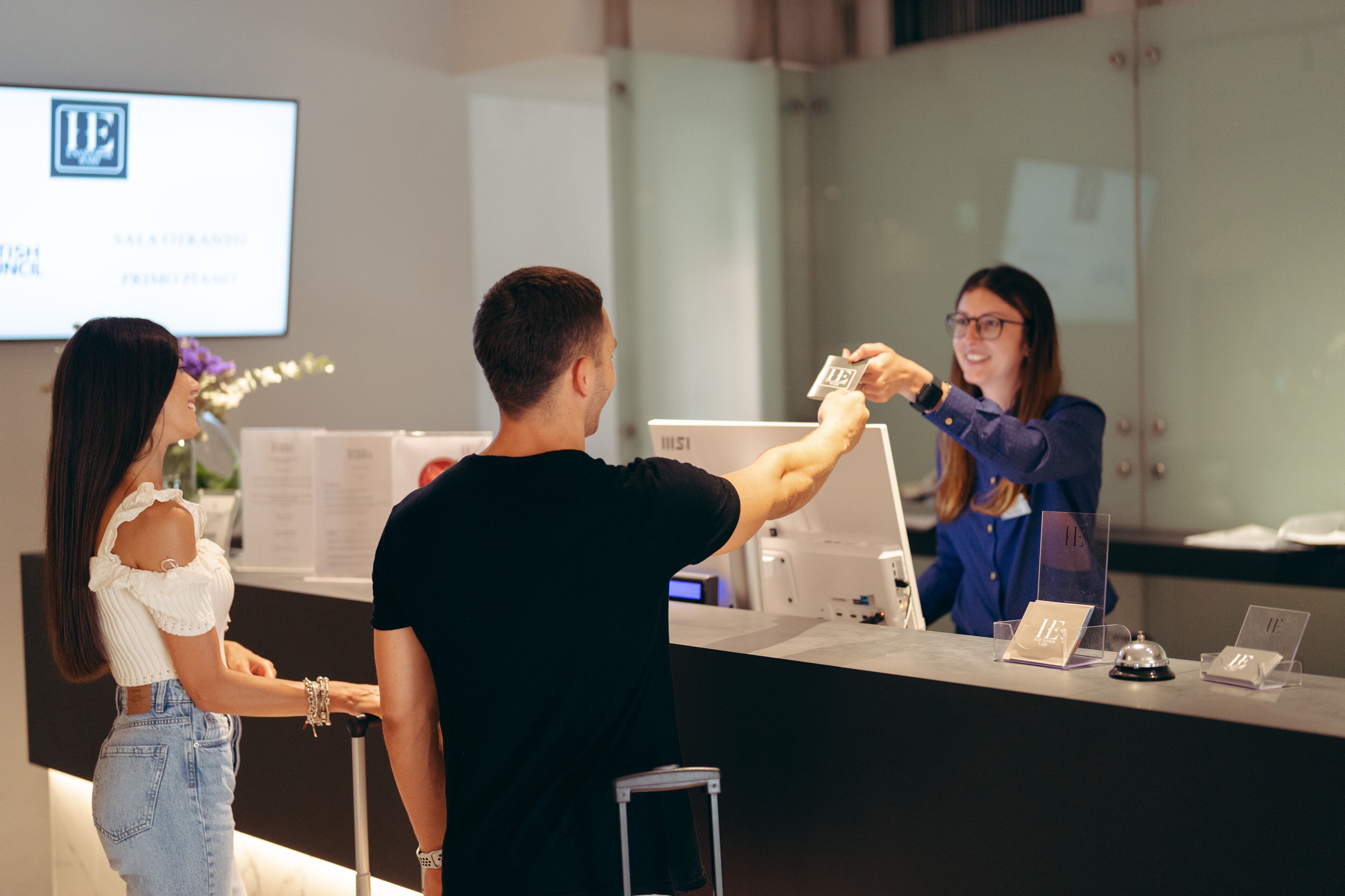 A reception desk with a digital screen displaying a logo and text, a glass partition, and a small table with a bell and name card holder.