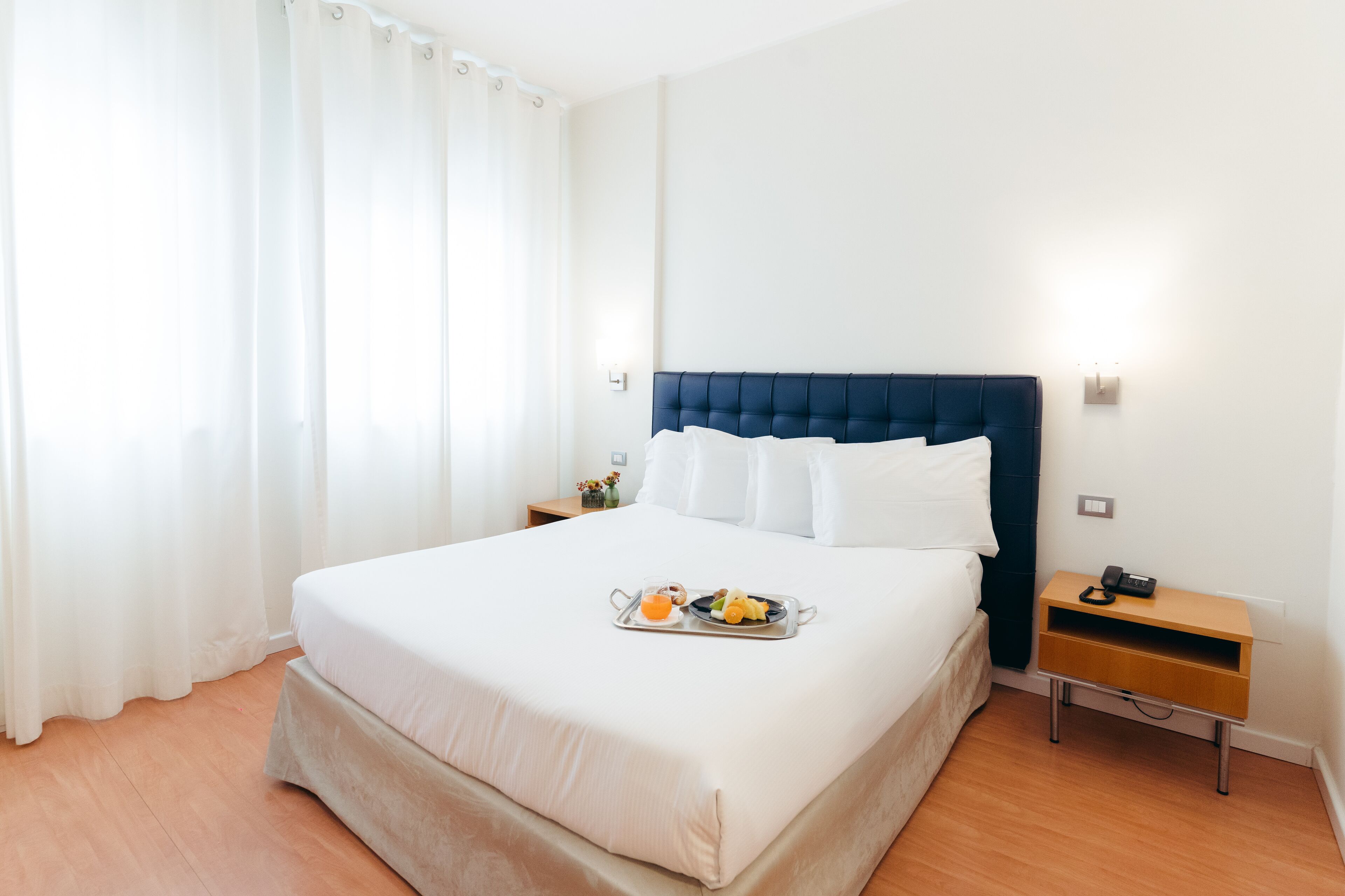 A neatly made bed with a tray of breakfast items, a bedside table with a lamp, and a window with curtains.