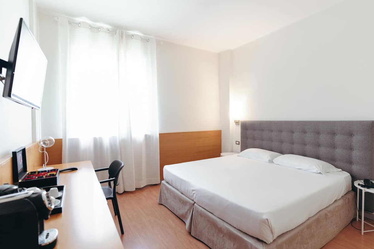 Ein Hotelzimmer mit Bett, Schreibtisch, Stuhl, Fernseher und einem Fenster mit Vorhängen.