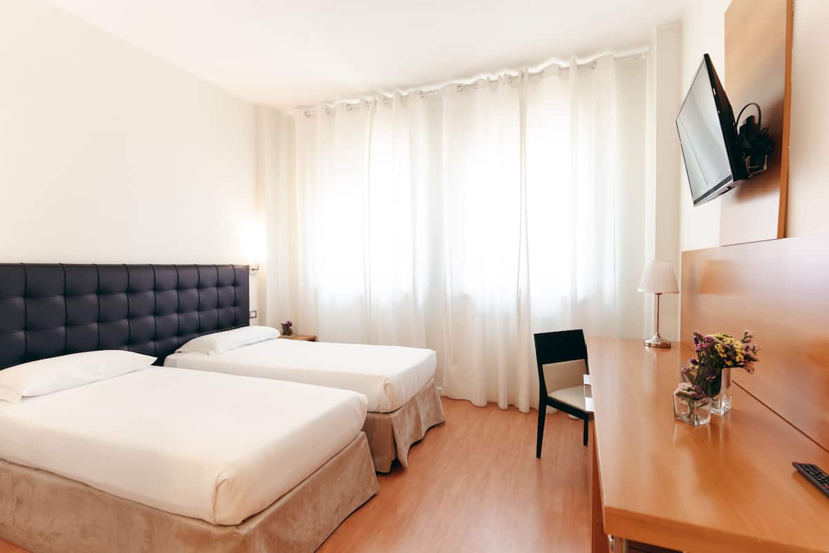 Ein Hotelzimmer mit zwei Betten, einem Schreibtisch, einem Stuhl, einem an der Wand befestigten Fernseher und einem Fenster mit Vorhängen.