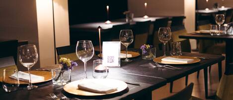 Un comedor con mesas dispuestas para dos, provistas de copas de vino, platos y cubiertos.