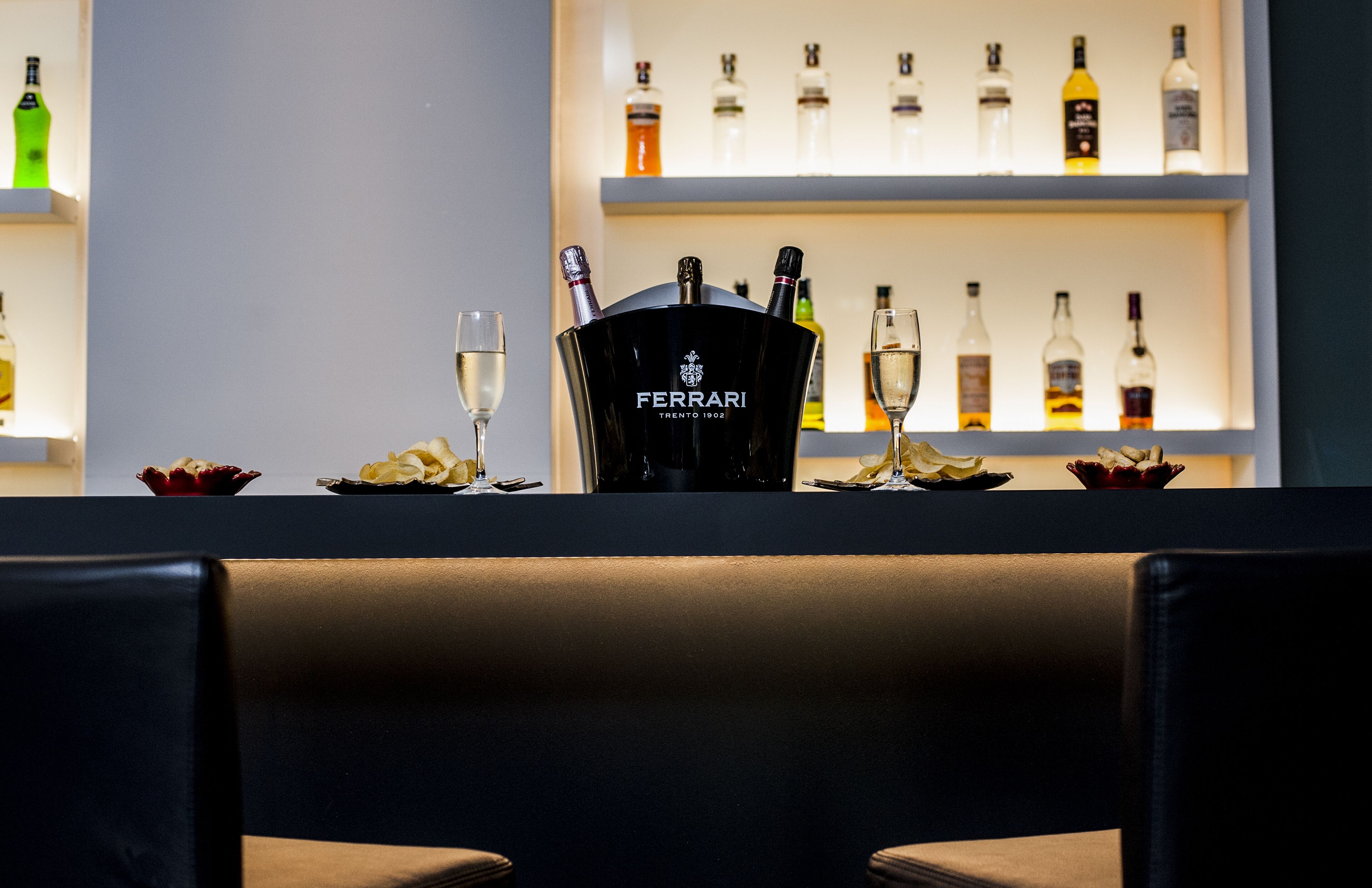 Een bar met een champagnekoeler met Ferrari-logo, glazen en een selectie drankflessen op planken.