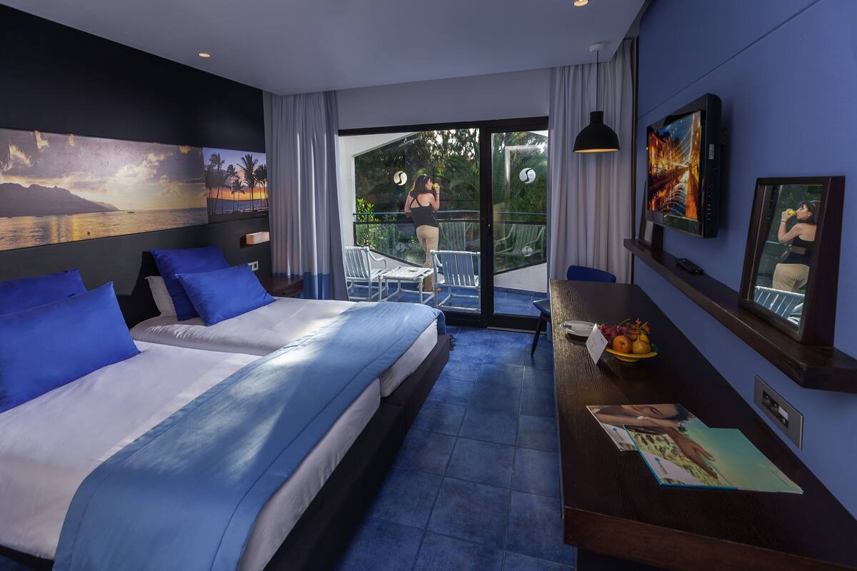 Habitación de hotel con una cama grande, un escritorio con computadora, un televisor y un balcón con vista al mar.