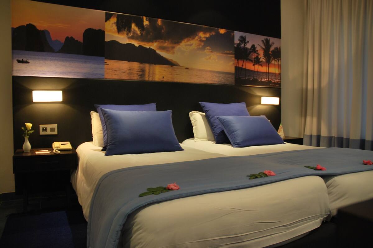 Habitación de hotel con una cama grande, dos mesitas de noche y un aparador, y una pared decorada con una pintura de atardecer.