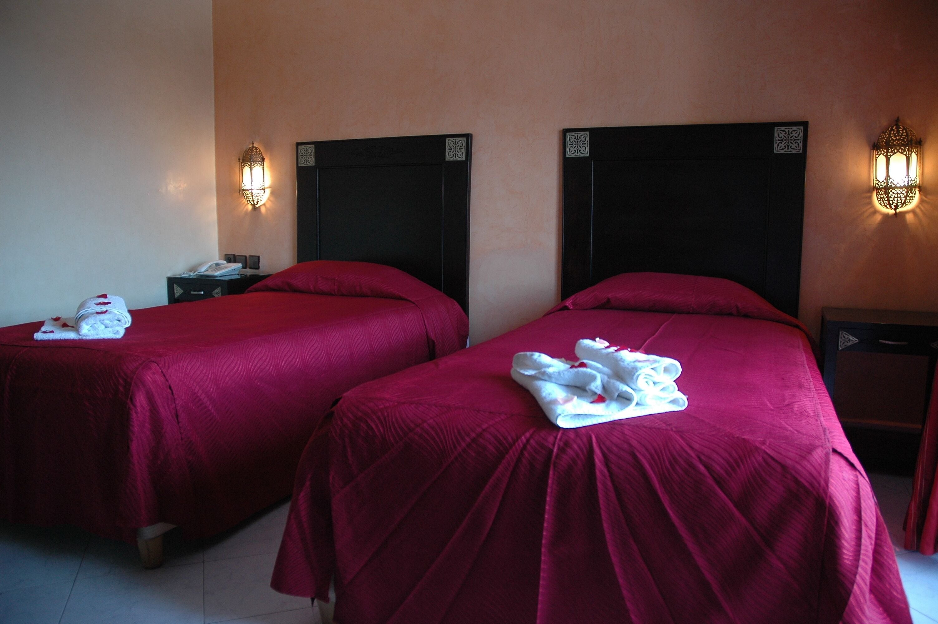 A hotel room with two beds, each with a red bedspread and a white towel on the bed. There are wall-mounted light fixtures on either side of the beds.