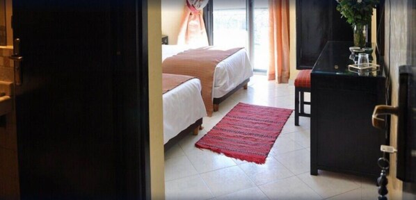 A hotel room with two beds, a red rug, a small table with a vase, and a view of the outdoors. - Hotel Mont Gueliz (Marrakech)