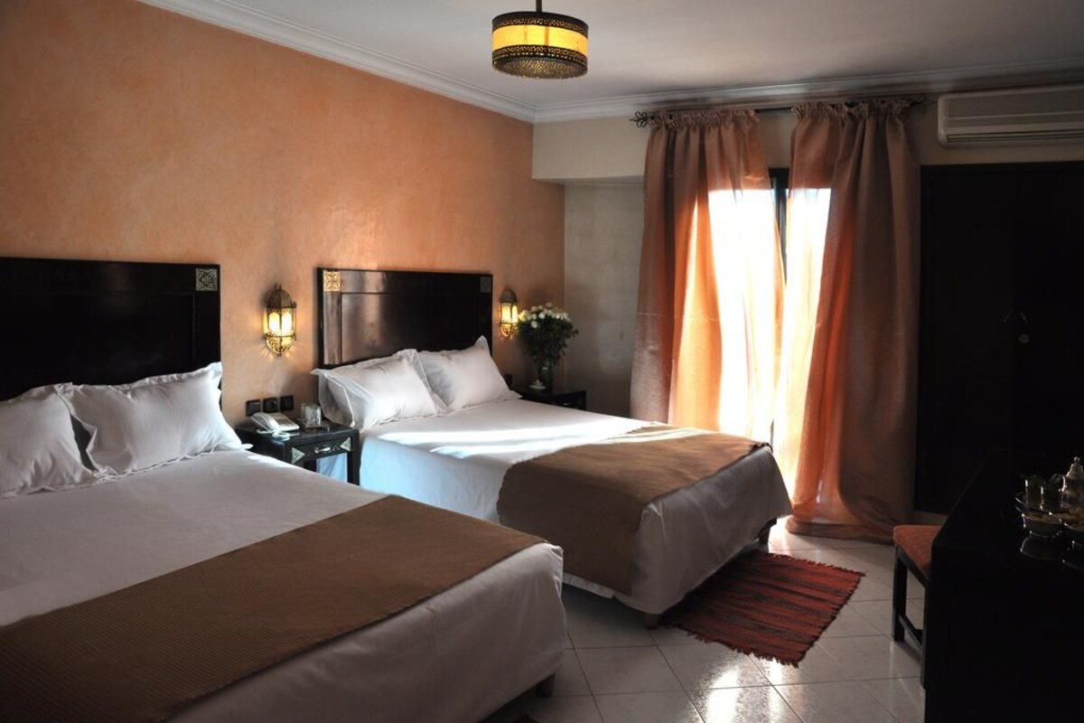 Une chambre d’hôtel avec deux lits, une table de chevet avec une lampe, un bureau avec une chaise et une grande fenêtre avec des rideaux.