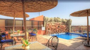A rooftop pool area with tables and chairs, surrounded by thatched umbrellas, and a rock wall. - Hotel Mont Gueliz (Marrakech)