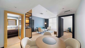 A modern hotel room with a wooden dining table, two armchairs, a sofa, a small kitchen area, and a large mirror.