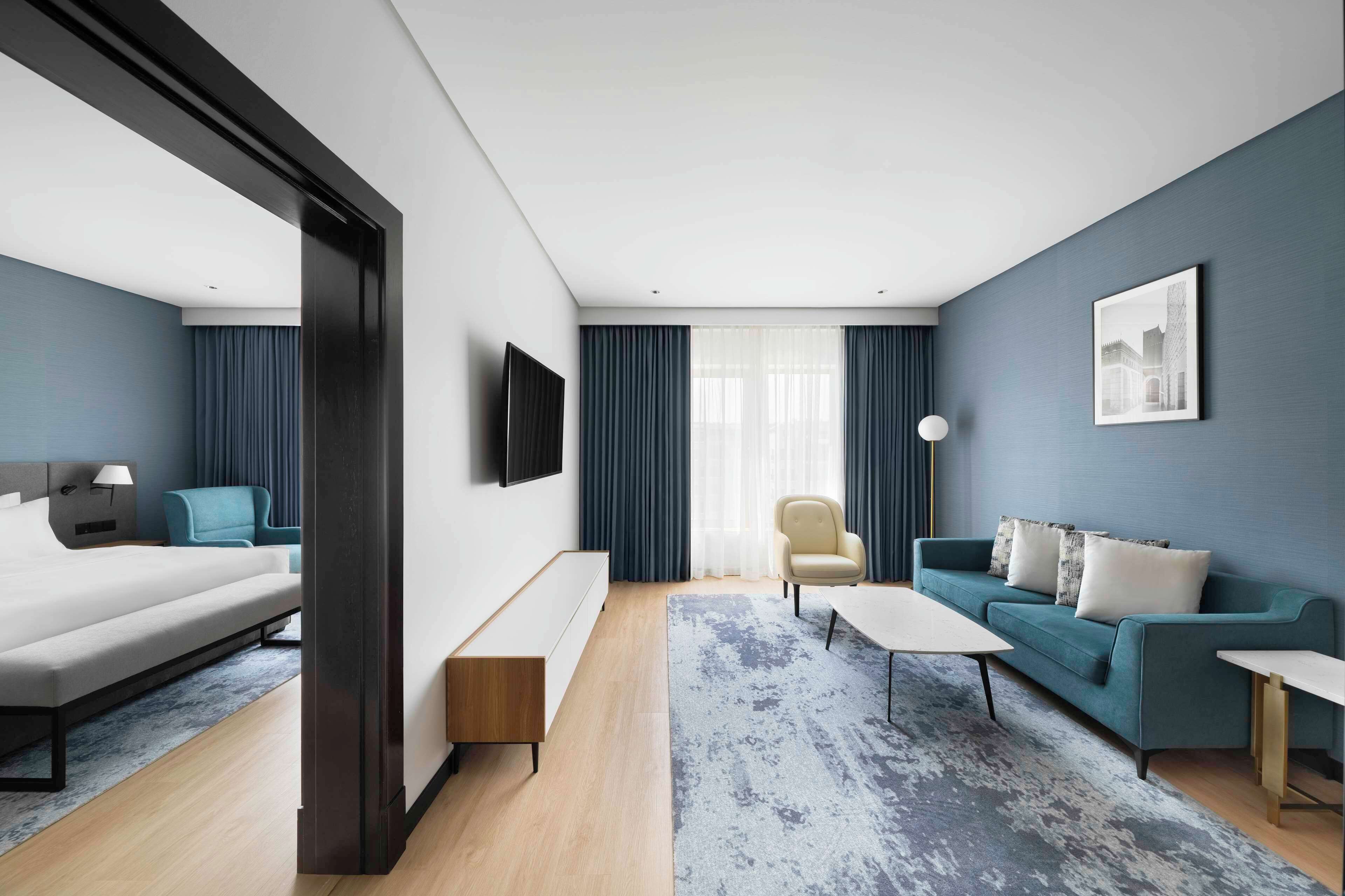 A modern hotel room with a bed, sofa, armchair, and a TV mounted on the wall.