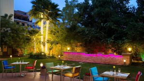 An outdoor dining area with multiple tables and chairs, surrounded by lush greenery and decorative lighting.