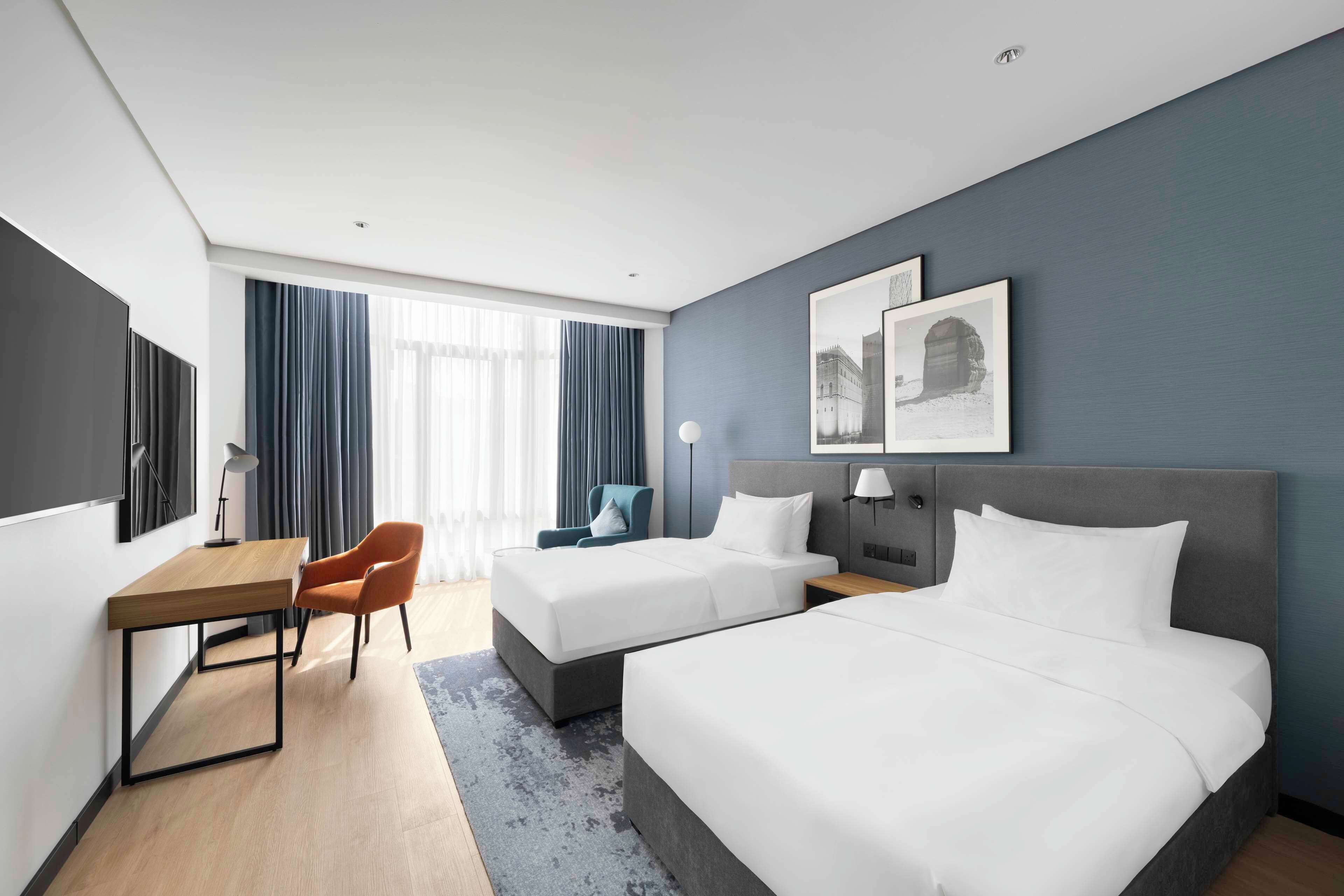 A hotel room with two beds, a desk, a chair, and a large window with curtains.
